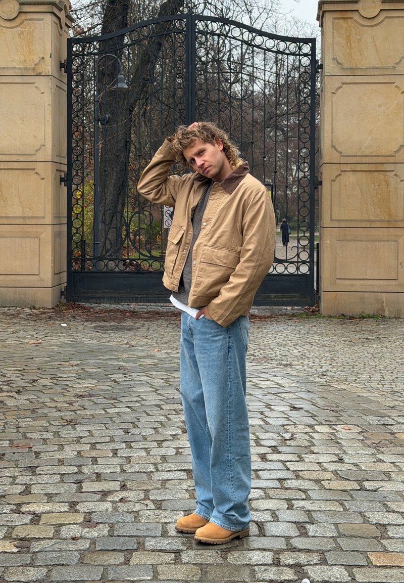 Veste matelassée beige avec col marron, pull en tricot gris, jean bleu clair et bottes beiges, debout près d'une porte en fer forgé ornée.