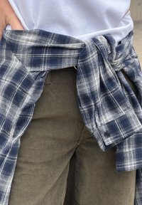 Cămașă din flanel în carouri albastre și albe legată în jurul taliei peste o cămașă albă, asortată cu pantaloni din catifea cu dungi verticale, de culoare verde măsliniu.