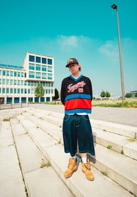 Černá mikina s červenými a modrými akcenty, džíny šortky a hnědé pracovní boty. Pozadí tvoří moderní vícepodlažní budova.