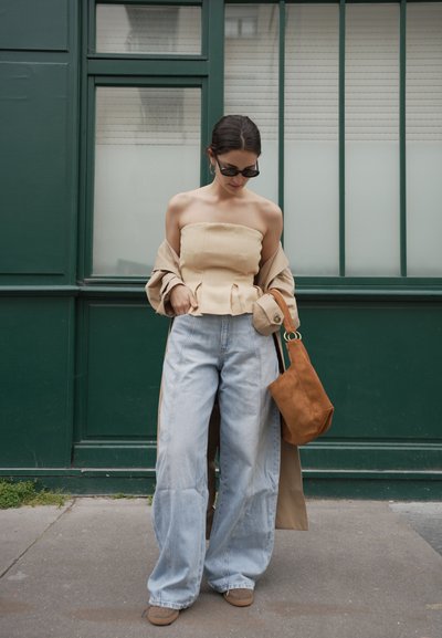 Mujer con top beige sin tirantes, vaqueros azul claro de gran tamaño, gafas de sol, abrigo marrón claro caído de los hombros y bolso marrón al hombro, de pie frente a una puerta verde.