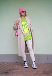 Μπεζ παλτό με διπλό κούμπωμα, φωτεινό πράσινο t-shirt με γραφικό σχέδιο λεμονιού, ανοιχτό ροζ κολάν, ροζ σκουφάκι, μικρή ροζ τσάντα, γυαλιά ηλίου.