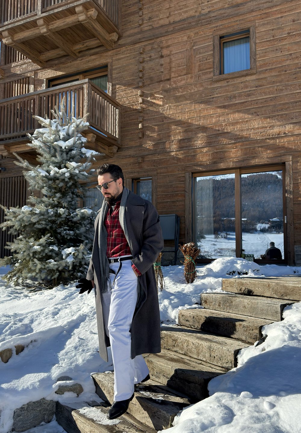 Homme portant des lunettes de soleil, un manteau gris, une chemise à carreaux rouges et un pantalon blanc descendant des escaliers en bois enneigés près d'un bâtiment en bois avec un pin.