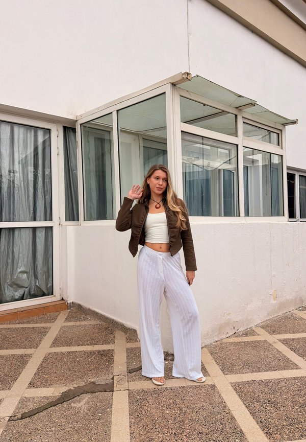 Jonge vrouw met lang haar, gekleed in een bruine jas, een wit crop top en losse witte broek, staand op een patroonachtige bestrating naast een wit gebouw.