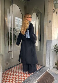 Casaco preto com mangas oversized, calças largas pretas e uma bolsa azul clara. O fundo apresenta azulejos padrão e portas de vidro.