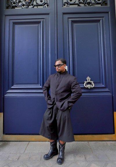 Persona vestida con un blazer negro oversized, pantalones cortos negros sueltos, botas negras y gafas de sol oscuras, de pie contra grandes puertas dobles azules ornamentadas.