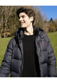 Tumši zils pūkains džemperis ar rāvējslēdzēju un kapuci, uz krūtīm sarkans logotips. Valkāts virs melnas pītās džempera, fonā zaļš ainavas skats.