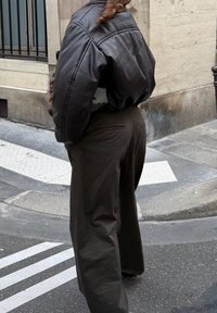 Une personne portant une veste en cuir noire et un pantalon ample de couleur sombre se tient sur un trottoir en ville près d'un bord de trottoir et d'un passage pour piétons.