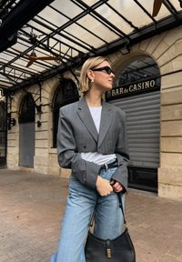 Donna che indossa un blazer grigio corto, camicia bianca, jeans azzurri chiari e occhiali da sole neri, mentre tiene una borsa nera fuori dal Bar du Casino.