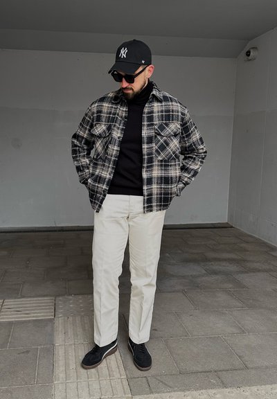 Hombre con chaqueta de cuadros en blanco y negro, cuello alto negro, pantalones blancos, zapatos negros, gafas de sol y gorra negra, de pie sobre un suelo de baldosas.