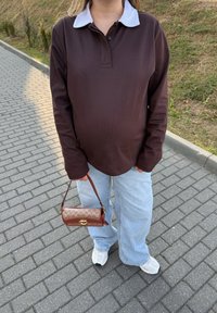Osoba w brązowej koszulce polo z długim rękawem, jasnoniebieskich dżinsach, białych trampkach, trzymająca małą brązową torebkę w wzory, stojąca na wybrukowanym chodniku.