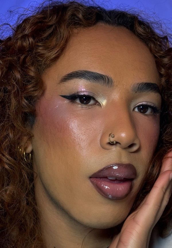 Retrato en primer plano de una persona con el pelo rizado, maquillaje de ojos brillante, pintalabios color ciruela con acabado gloss, un aro en la nariz y una mano apoyada en la mejilla.