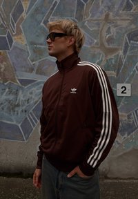Brązowa kurtka zapinana na zamek z białymi paskami na rękawach, z logo Adidas. Noszona z jasnoniebieskimi dżinsami na tle teksturowego muralu.