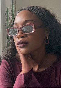 Lyserøde rektangulære solbriller med mørke linser, båret af en person med flettet hår. Baggrunden viser grønt løv, hvilket tilføjer dybde til scenen.