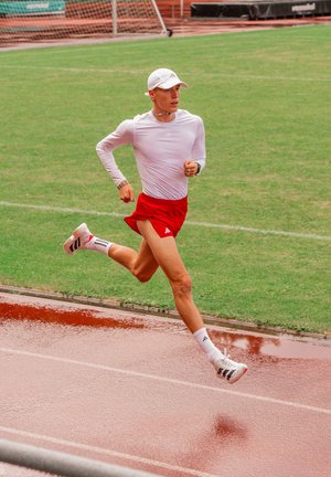 Runner wearing white long-sleeve shirt, red shorts, and white cap sprinting on wet outdoor track beside green field.