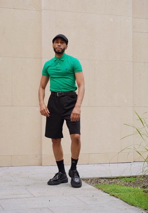 Camisa polo verde, pantalones cortos negros, cinturón negro, zapatos negros, calcetas negras con patrón y gorra negra. De pie frente a una pared clara con vegetación.