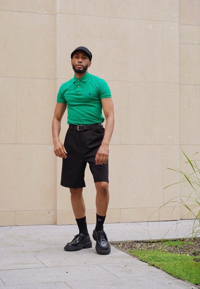 Camisa polo verde, pantalones cortos negros, cinturón negro, zapatos negros, calcetas negras con patrón y gorra negra. De pie frente a una pared clara con vegetación.