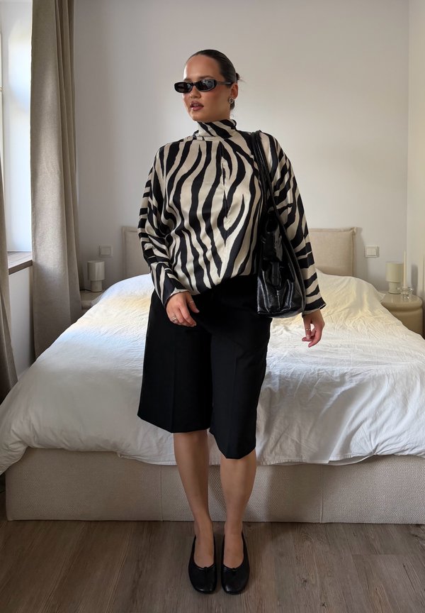 Femme portant des lunettes de soleil noires, un col roulé imprimé zèbre, un short noir mi-long et tenant un sac noir, debout dans une chambre.