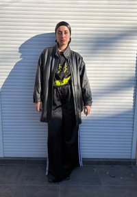 Personne portant une veste en cuir noire, un t-shirt graphique noir, un pantalon large noir avec des rayures blanches, et un foulard noir, debout devant une porte roulante blanche.