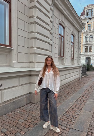 Camisa blanca de manga larga y botones con un brillo sutil, combinada con jeans oscuros de pierna ancha y zapatillas ligeras, de pie en una calle empedrada.