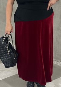 Burgundi bársony maxi szoknya légies szabással, fekete szűk felsővel párosítva. Egy texturált fekete kézitáska ezüst láncpánttal van a kezében.