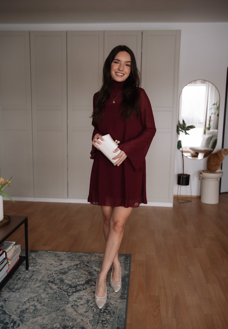 Γυναίκα σε μακρυμάνικο μπορντό φόρεμα που κρατά ένα λευκό τσαντάκι, στέκεται σε χαλί σε ένα μοντέρνο σαλόνι με έναν καθρέφτη και μια γάτα κοντά.