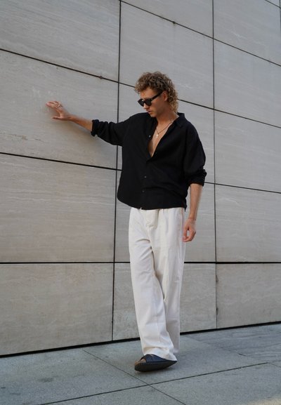 Camisa negra de botones, pantalones anchos blancos y zapatos oscuros. Pelo rizado, complementado con gafas de sol y un collar con una cruz. Fondo de pared texturizada.