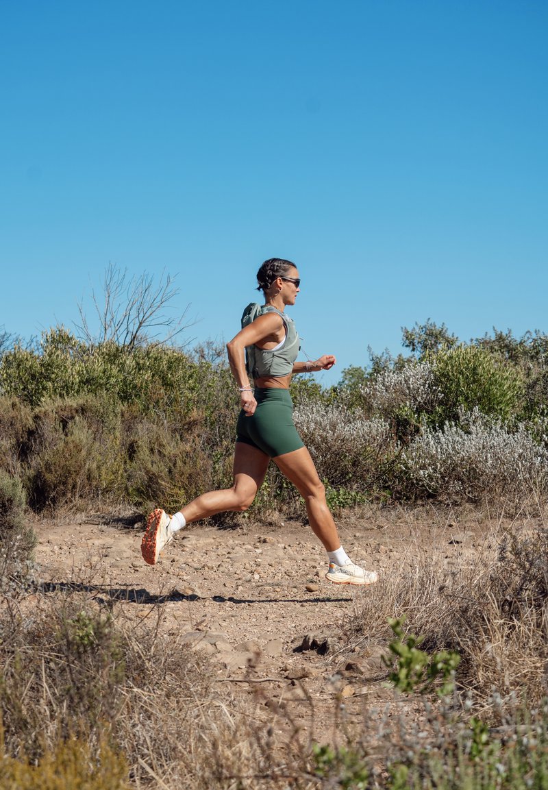 Femme en tenue de sport courant sur un sentier sec et rocailleux entouré d'arbustes sous un ciel bleu clair.