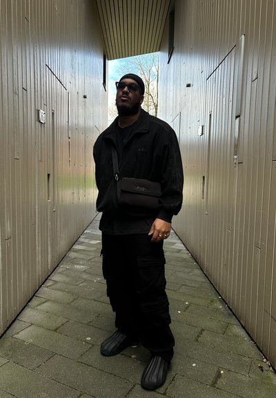 Chaqueta negra con cremallera y rayas laterales, camiseta negra, pantalones cargo y zapatos de lona texturizados. De pie en un pasillo estrecho con paneles de madera.