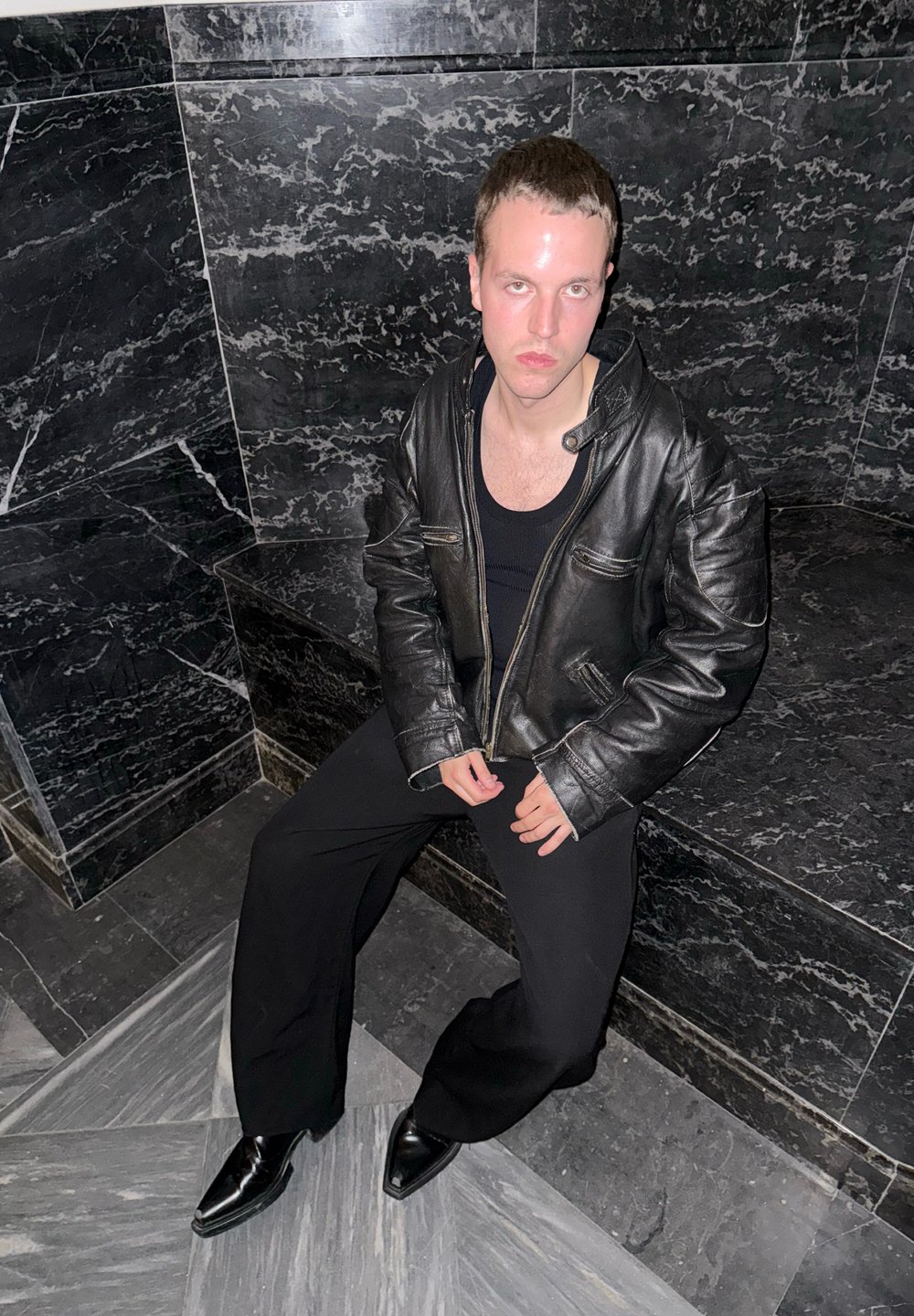 Musta nahkatakki vetoketjulla ja kahdella rintataskulla, yhdistetty tiukkaan mustaan toppiin ja leveisiin mustiin housuihin. Kiiltävät mustat kengät.