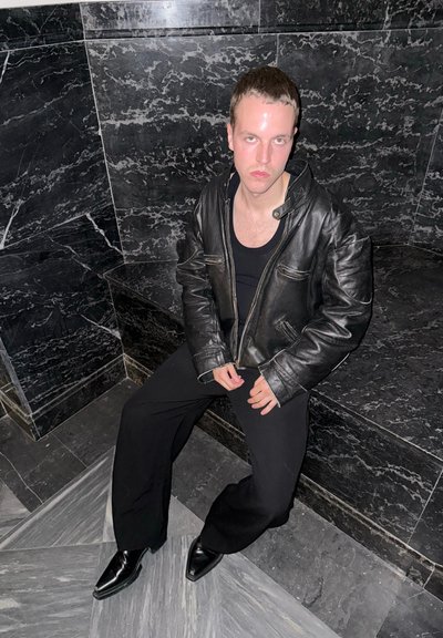 Chaqueta de cuero negra con cierre de cremallera y dos bolsillos en el pecho, combinada con una camiseta negra ajustada y pantalones anchos negros. Zapatos negros brillantes.