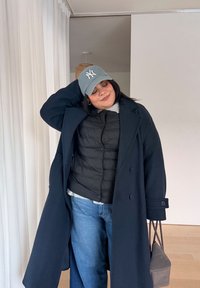 Donna con cappellino da baseball blu, cappotto lungo scuro, giacca imbottita nera e jeans, in piedi all'interno, che tiene una borsa grigia, con una mano dietro la testa.