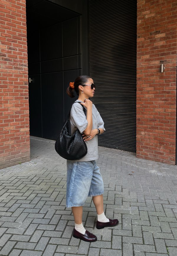 Woman in sunglasses and gray shirt stands on sidewalk holding black bag, with red brick walls and black doors in the background.