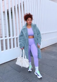Femme aux cheveux bouclés portant un manteau bleu duveteux, des leggings lavande et un crop top, des baskets blanches, et portant un grand sac blanc, debout près d'une clôture blanche.