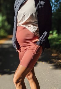 Dulkesnės rožinės dviračių šortai su aukštu liemeniu ir šoninėmis kišenėmis, derinami su kreminės spalvos marškiniais ir tamsia striuke, fone – natūrali aplinka.