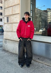 Κόκκινο μπουφάν με άσπρο λογότυπο, μαύρη κουκούλα από κάτω, φαρδιά σκούρα τζιν παντελόνια και μαύρα παπούτσια. Στέκεται δίπλα σε τοίχο από πέτρα και παράθυρο.