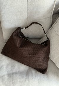 Brązowa pleciona skórzana torba o trapezoidalnym kształcie, z zamkiem błyskawicznym i plecioną rączką, spoczywająca na jasnym materiale.