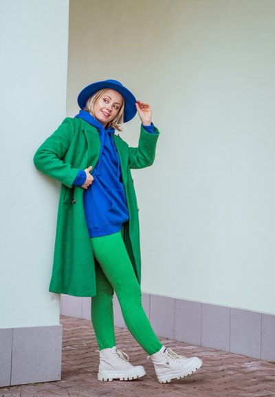 Abrigo de lana verde, sudadera azul, leggins verdes y zapatillas gruesas blancas. Un sombrero azul y un fondo de pared texturizada completan el look.