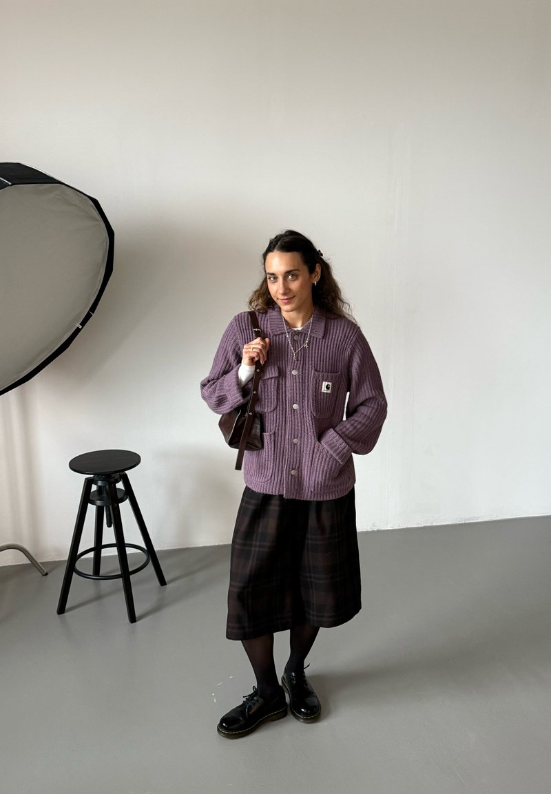 Kvinna med mörkt hår bär en lila stickad jacka, svart rutig kjol och svarta skor, står i en studio med en pall och en softbox-lampa i närheten.