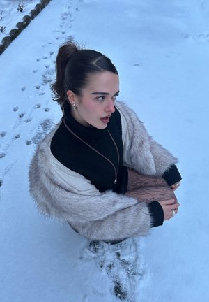 Joven mujer con suéter negro y abrigo de piel sentada en la nieve, mirando hacia un lado, con huellas visibles en el camino nevado cercano.