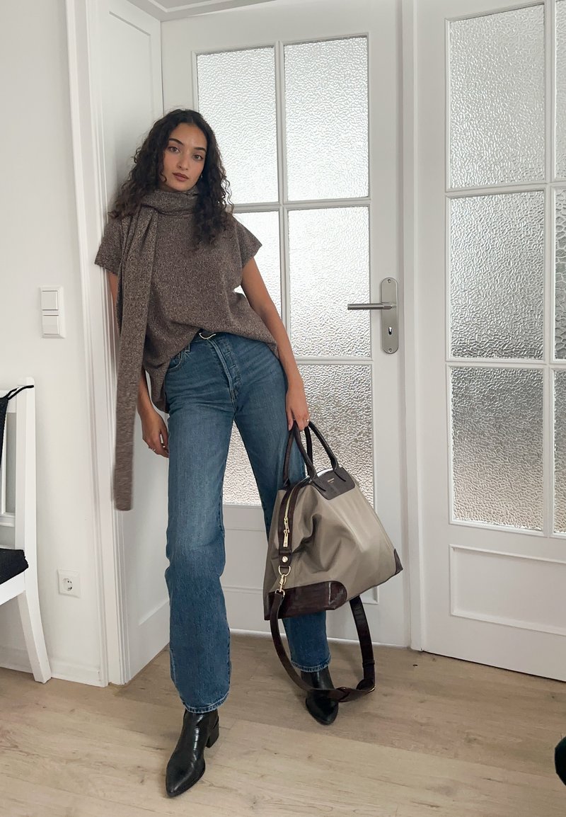 Haut en maille gris-brun avec un détail écharpe, jean évasé bleu clair, et bottines noires. Porte un grand sac gris avec des accents sombres.