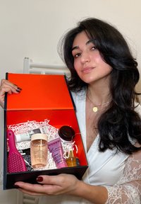 Giovane donna che tiene in mano una scatola regalo aperta con prodotti per la cura dei capelli e una spazzola per capelli rosa all'interno, indossando una vestaglia di pizzo bianco.