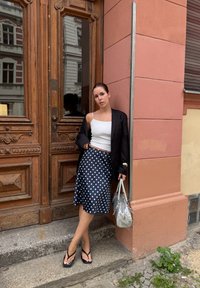 Čierny blazer cez biely tielko, midi sukňa s bodkami, čierne žabky a textúrovaná strieborná kabelka na pozadí drevených dverí.