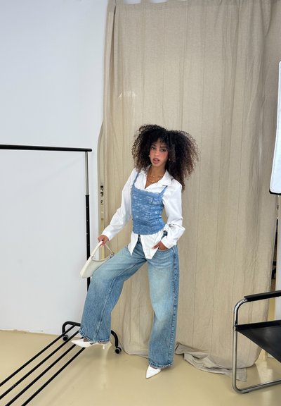 Corsé de encaje azul sobre una camisa blanca, combinado con jeans de pierna ancha deslavados y tacones blancos puntiagudos. La modelo sostiene un pequeño bolso blanco.