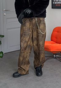 Personne portant des gants noirs, des bottes noires, un pantalon large brun vieilli avec une chaîne, debout près d'une chaise orange et d'une porte blanche.