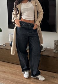 Jean bleu foncé à jambes larges et taille haute, avec des coutures apparentes. Associé à un haut blanc ajusté et un trench-coat beige. Sneakers blanches.