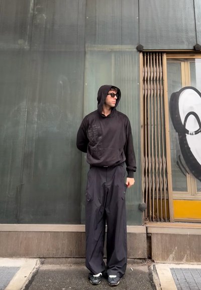 Sudadera negra con corte holgado y bolsillo frontal, combinada con pantalones anchos negros con bolsillos laterales. Unas gafas de sol completan el conjunto.