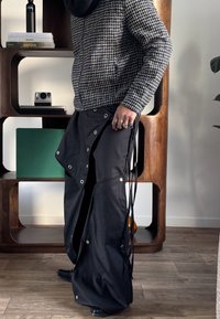 Personne portant une veste à carreaux noir et blanc et un pantalon large noir à boutons pression, debout à l'intérieur près d'une étagère en bois.