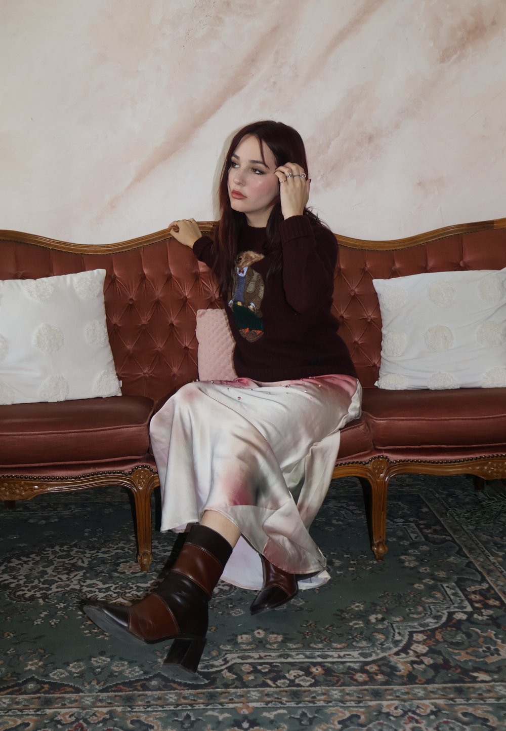 Sweter dzianinowy z motywem zwierzęcym, pastelowa satynowa spódnica oraz brązowe botki na obcasie. Usytuowane na pikowanej różowej aksamitnej sofie z dekoracyjnymi poduszkami.