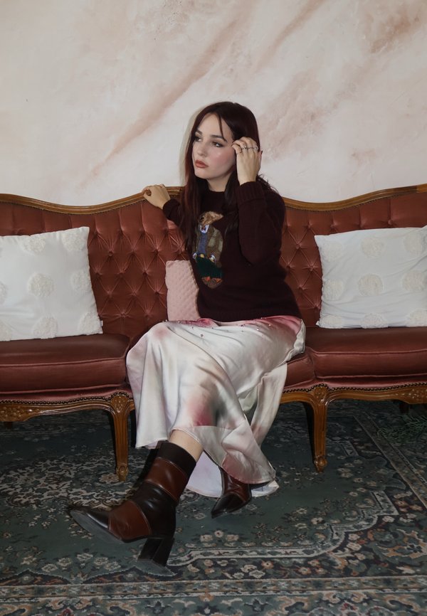 Knit sweater with an animal motif, pastel satin skirt, and brown heeled boots. Set on a tufted pink velvet sofa with decorative pillows.