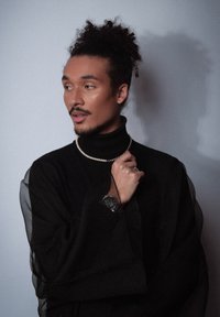 Pull noir à col roulé avec des manches transparentes, orné d'un collier de perles. Portant une montre, le sujet a les cheveux bouclés et une expression pensive.